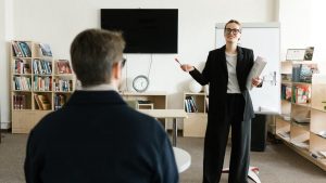 A teacher in a black blazer conducts a session with a student in a library setting.