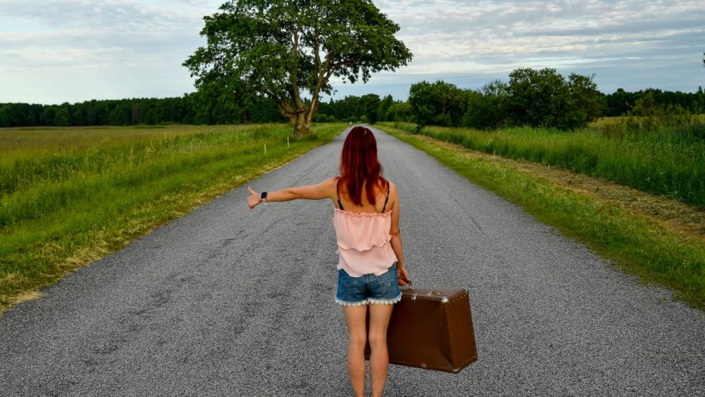 Red-haired woman hitchhiking with vintage suitcase on scenic rural Estonian road. Summer adventure.